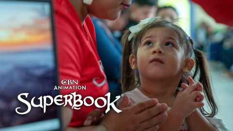 A little girl in Honduras folds her hands to pray and looks up at the Superbook volunteer next to her. The CBN Animation and Superbook logos are in the bottom left corner of the photo.