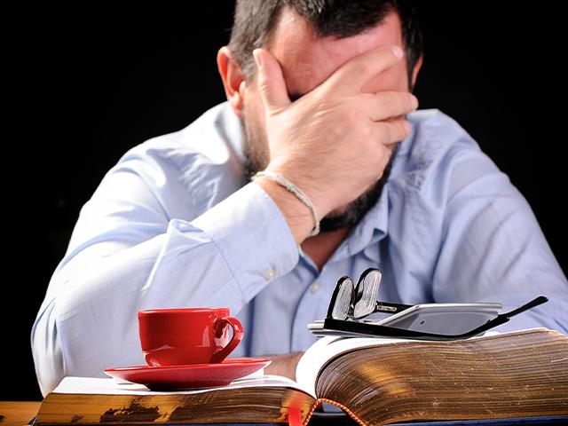 man sitting at desk with coffee cup on an open book and head in his hands