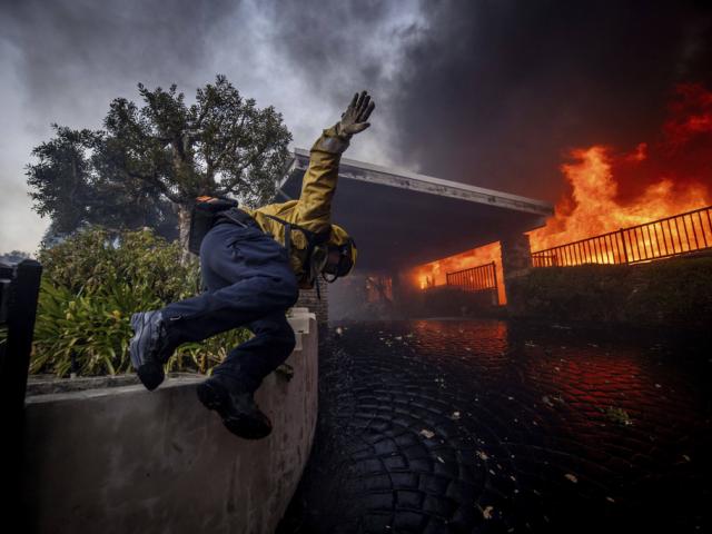 A firefighter jumps over a fence while fighting the Palisades Fire in the Pacific Palisades neighborhood of Los Angeles, Tuesday, Jan. 7, 2025. (AP Photo/Ethan Swope)