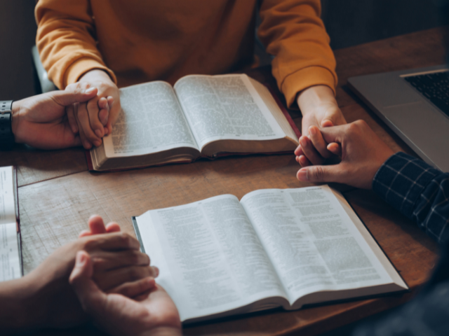 two open Bibles on a study table with people holding hands
