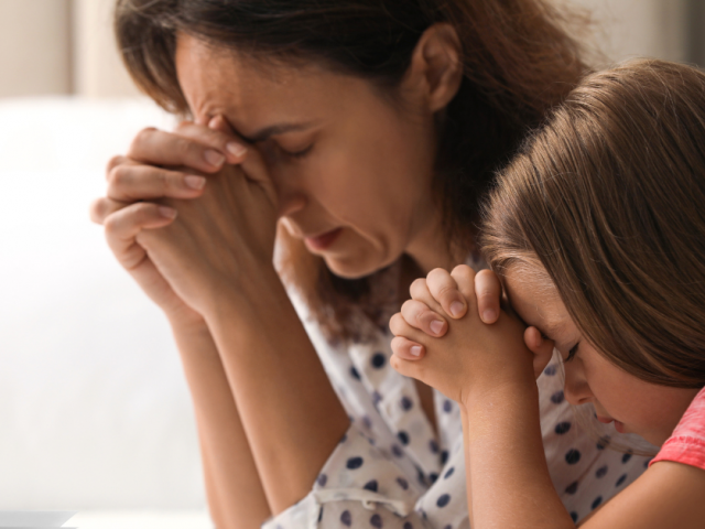 a mother and young daughter kneeling in prayer on a bedside