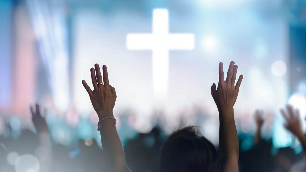 Christian churchgoers raise their hands in worship to Jesus as a cross shines on stage. (Adobe stock image)